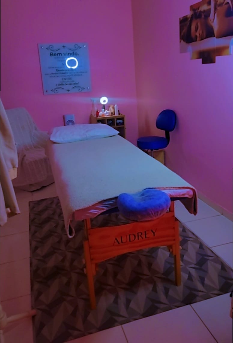 Sala de massagem vazia com paredes rosa, maca de madeira Audrey, apoio de rosto azul e placa de boas-vindas