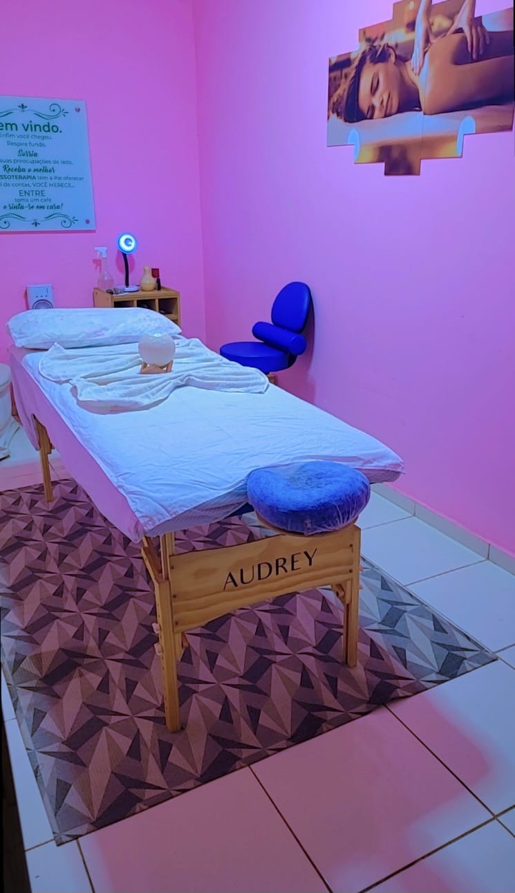 Ângulo da maca Audrey com toalhas, luminária de sal, cadeira azul e quadro de massagem na parede